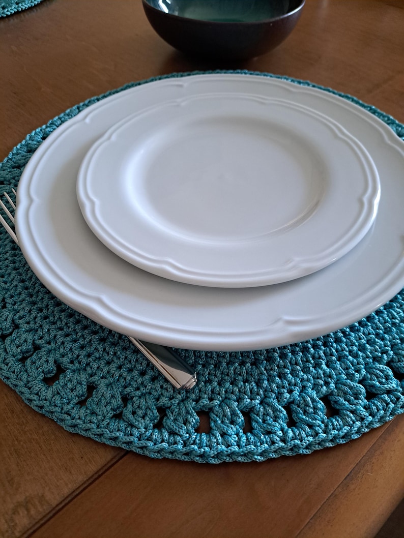 May include: A teal crocheted placemat with a white dinner plate and a smaller white plate on top. A silver fork and knife are placed on the placemat.