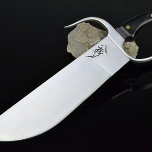 May include: A silver-colored Kung Fu sword with a black handle and gold accents. The sword has a curved blade and a ring handle.