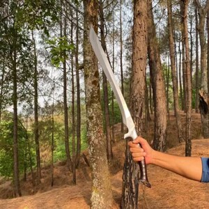 May include: A silver sword with a brown leather handle, held by a person's hand in a forest setting.