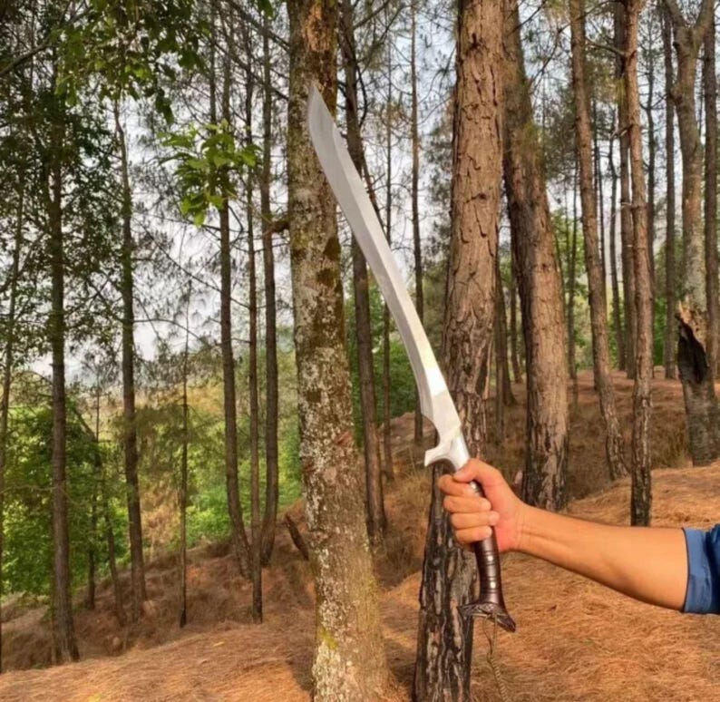 May include: A silver sword with a brown leather handle, held by a person's hand in a forest setting.