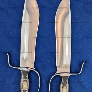 Peut inclure: Deux grandes &eacute;p&eacute;es en acier poli avec des lames incurv&eacute;es et des poign&eacute;es distinctives. Les poign&eacute;es pr&eacute;sentent un motif de grain de bois fonc&eacute; avec un design ovale central. Les &eacute;p&eacute;es sont pr&eacute;sent&eacute;es sur fond bleu.