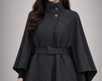 May include: A charcoal gray cape coat with a high collar and a matching belt. The coat features a button closure and a flared silhouette. The model is wearing gold hoop earrings and a black sweater underneath.