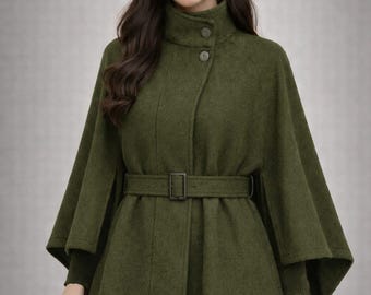 May include: Olive green cape coat with a high collar and a matching belt. The coat features a button closure and a flared silhouette. The model is wearing gold hoop earrings and a dark green sweater underneath.
