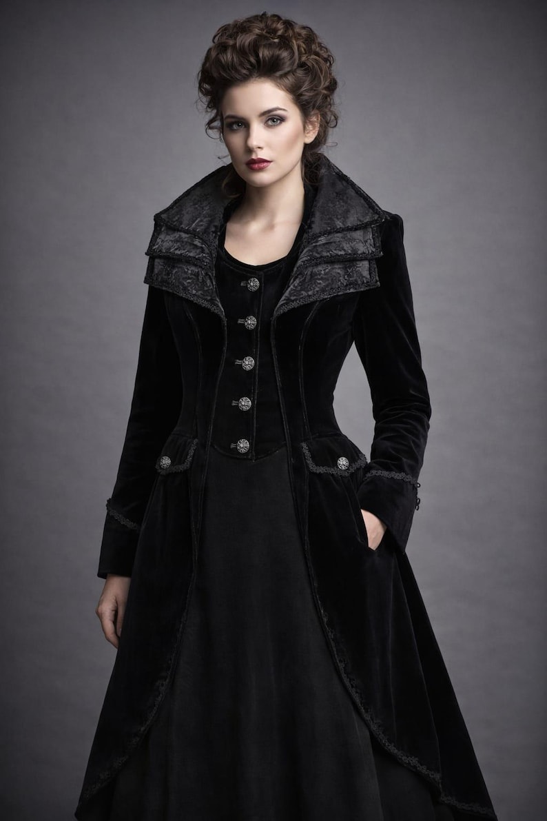 May include: A long, black velvet coat with a high collar and decorative buttons. The coat features intricate detailing on the collar and pockets, and is worn over a black dress. The overall style is gothic and elegant.