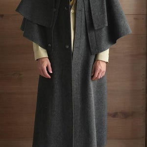 May include: A dark gray wool coat with a cape-like design, featuring a fur collar and a button-down front. The coat is paired with a light-colored shirt, a striped tie, and dark pants. The image includes the text "CLOTHING BY MAAZ".