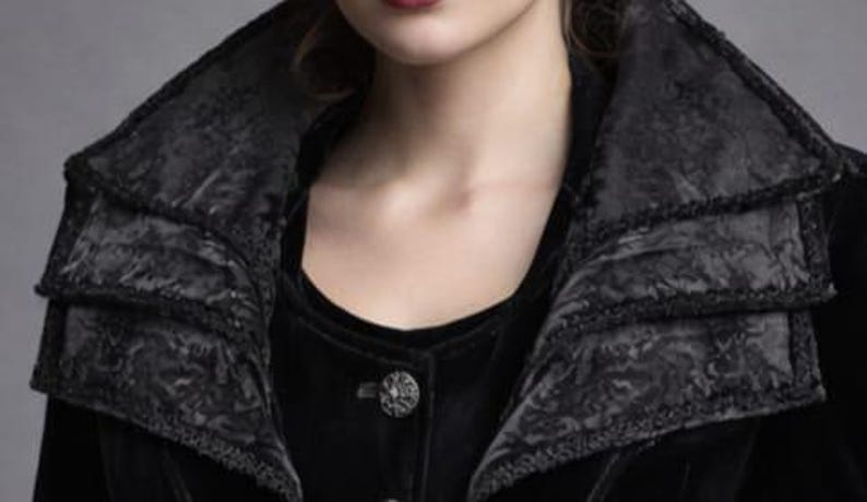 May include: A black velvet jacket with a layered collar and ornate button. The collar has a textured, patterned design. The jacket is buttoned up, and the fabric appears soft and luxurious. The overall style suggests a gothic or vintage aesthetic.