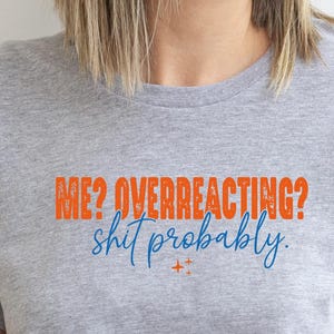 May include: A heather gray short-sleeve t-shirt featuring the orange text ME? OVERREACTING? and the blue cursive text shit probably. The shirt has a crew neckline and rolled sleeves, with a small orange star icon below the blue text.