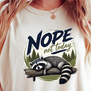 May include: An ivory t-shirt featuring a graphic of a raccoon lying on a tree branch. The text NOPE not today is printed above the raccoon in dark blue and green. The design includes a circular background with green pine tree silhouettes.