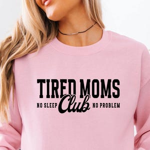 May include: A light pink long-sleeve sweatshirt featuring black text that reads TIRED MOMS CLUB with the words NO SLEEP and NO PROBLEM flanking the word Club. The garment has a crew neckline and ribbed cuffs, providing a simple and clean look.