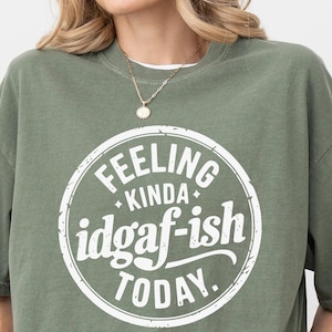 May include: A moss green short-sleeve t-shirt featuring a white circular graphic on the chest. The text inside the circle reads "FEELING KINDA idgaf-ish TODAY." with small star accents. The shirt has a crew neckline and a relaxed, loose fit.