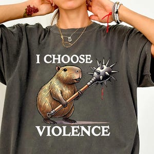 May include: Dark grey t-shirt with the text "I Choose Violence" and a cartoon capybara holding a spiked mace. The capybara is brown and tan, and the mace has silver spikes and a wooden handle. The shirt is a Pepper color.