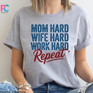 May include: A light gray t-shirt with the text "MOM HARD, WIFE HARD, WORK HARD, Repeat" in blue and red lettering. The shirt is made of a soft material and is suitable for casual wear.