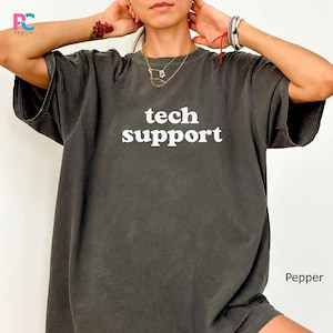 May include: A dark gray t-shirt with the words "tech support" in white block letters. The shirt has short sleeves and a relaxed fit. The word "Pepper" is printed in the bottom right corner.