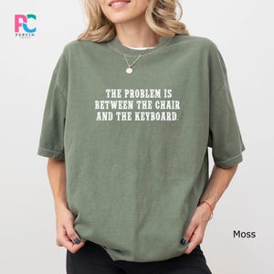 May include: A moss green t-shirt with the text "THE PROBLEM IS BETWEEN THE CHAIR AND THE KEYBOARD" in white. The shirt is a casual style, with short sleeves and a relaxed fit. The shirt is made of a soft material.