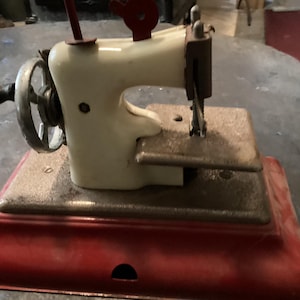 May include: A vintage-style toy sewing machine featuring a cream-coloured body, a red base, and a silver hand crank with a black handle. The machine includes a textured grey metal platform, a vertical needle bar, and a small red lever on top.
