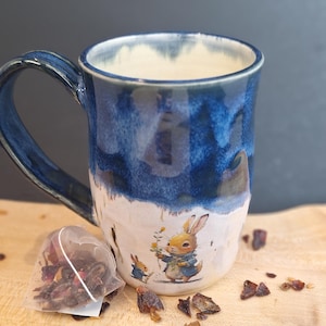 Peut inclure: Une tasse en céramique avec un glaçage bleu et blanc, ornée d'une illustration de lapin. Un sachet de thé et des feuilles de thé en vrac sont à côté de la tasse sur une surface en bois. La tasse a une grande anse.
