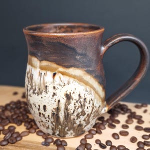 May include: A ceramic mug with a curved handle, featuring a two-tone glaze. The upper portion is a dark, solid brown, while the lower section is a cream colour with dark brown, tree-like drip patterns and speckles throughout the surface.