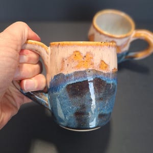 May include: Two ceramic mugs with a blue, brown and orange glaze. The mugs have a rounded shape and a handle.