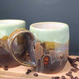 Peut inclure: Deux tasses en céramique avec un émail réactif présentant une partie supérieure vert clair qui se fond dans une base brun foncé. Les tasses ont une forme arrondie avec une anse incurvée et un motif de coulures où les couleurs se mélangent.