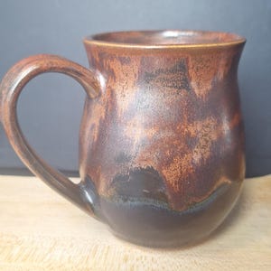 May include: A ceramic mug with a rounded body and a curved handle. The exterior features a mottled glaze with blended tones of deep brown, copper, and black, creating a textured appearance. The rim is smooth and slightly flared outward from the body.