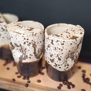 May include: Two ceramic travel mugs with a speckled cream upper section and a solid dark brown base. Each mug features a unique, indented grip area on the side and a semi-circular lid with a small drinking hole. The mugs have a textured, rustic finish.