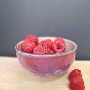 May include: A small, handmade ceramic bowl filled with fresh red raspberries. The bowl has a reddish-purple glaze with blue accents around the rim. Three raspberries sit outside the bowl on a wooden surface. The background is a dark grey.