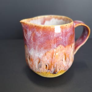 May include: A handmade ceramic jug with a pink, white and yellow glaze. The jug has a rounded body and a handle.