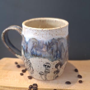 May include: A ceramic mug with a cream rim and a blue and grey glazed exterior. The mug features hand-drawn mushroom illustrations. Coffee beans are scattered on the wooden surface. The mug has a large handle.