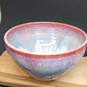 May include: A ceramic bowl with a smooth, glossy finish featuring a gradient of colours. The rim is a vibrant pinkish-red that transitions into a soft, pale blue and white interior. The base of the bowl is a neutral, unglazed off-white ceramic material.