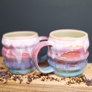 Peut inclure: Deux tasses en céramique présentant un corps arrondi et nervuré avec un émail multicolore. Les couleurs passent d'un goutte-à-goutte blanc crème sur le bord à des couches de rose, orange, violet et bleu. Chaque tasse a une anse incurvée.