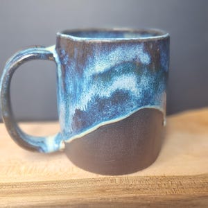 May include: A ceramic mug with a cylindrical body and a curved handle. The lower portion of the mug features a matte dark brown finish, while the upper portion is glazed in a vibrant, mottled blue and black pattern that drips downwards.