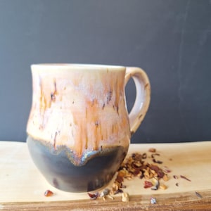 Peut inclure: Une tasse en céramique faite à la main avec un glaçage unique. La tasse présente une base sombre qui se transforme en nuances d'orange, de crème et de marron. La tasse mesure environ 10 cm de haut et possède une anse incurvée. Des feuilles de thé sont éparpillées sur une surface en bois.