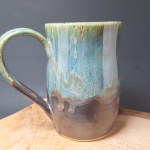 Peut inclure: Une tasse en céramique faite à la main avec un glaçage unique. La tasse présente une base marron foncé qui se transforme en nuances de bleu, vert et blanc. La poignée est d'une couleur complémentaire au corps de la tasse. La tasse est posée sur une surface en bois.