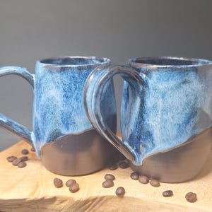 May include: Two handmade ceramic mugs with a blue and black glaze. The mugs have large handles and sit on a wooden board with scattered coffee beans. The mugs are approximately 10 cm tall.