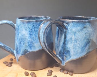 May include: Two handmade ceramic mugs with a blue and black glaze. The mugs have large handles and sit on a wooden board with scattered coffee beans. The mugs are approximately 4 inches tall.