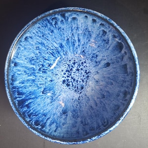 May include: A circular ceramic bowl with a deep blue glaze. The interior has a textured, swirling pattern of lighter blue and white, creating depth. The bowl's rim is a darker shade of blue.