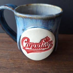 Peut inclure: Tasse en céramique bleue avec une anse. La tasse présente un emblème blanc avec le mot "Canadian" en rouge et une feuille d'érable rouge. La tasse a une finition brillante et repose sur une surface en bois.