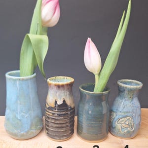 May include: Four small ceramic bud vases with unique glazes. Vase 1 is light blue with a tulip. Vase 2 has horizontal ridges and a dark brown and tan glaze. Vase 3 is dark blue-green. Vase 4 is light blue with a bee relief and a tulip.