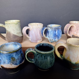 May include: A collection of seven small, handmade ceramic mugs in various colors and patterns. The mugs are all different shapes and sizes, and they have a rustic, handmade look.