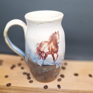 Peut inclure: Une tasse en céramique avec une partie supérieure blanche et une partie inférieure brune texturée. La tasse présente une illustration peinte d'un cheval brun à la crinière et à la queue fluides, debout dans un lavis bleu clair et blanc.