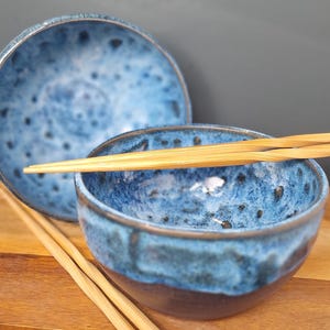 May include: Two ceramic bowls with a mottled blue and black glaze finish. The bowls feature a rounded shape with a dark base and a textured, speckled blue interior. A pair of light-coloured bamboo chopsticks rests across the top of one bowl.