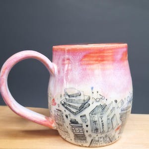 May include: A ceramic mug with a rounded body and a curved handle. The upper portion features a vibrant pink and orange gradient glaze, while the lower section is cream-coloured and decorated with black line drawings of stacked books and small hearts.