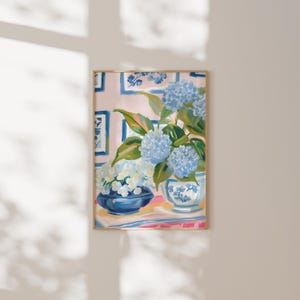 Pode incluir: Pintura emoldurada de hortênsias azuis e brancas em vasos decorativos. A obra apresenta um fundo rosa claro com detalhes florais azuis. As hortênsias estão numa tigela azul e num vaso branco com desenhos florais azuis.