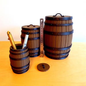 Puede incluir: Tres barriles de madera con bandas y tapas de metal. El barril más grande es marrón con una tapa negra. El barril del medio es marrón con una tapa negra. El barril más pequeño es marrón con una tapa negra y tiene dos bolígrafos dentro.