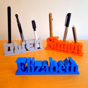 May include: Three 3D printed pen holders, each with a different name. The first is grey and says "Owen", the second is orange and says "Simon", and the third is blue and says "Elizabeth".