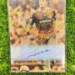 Könnte beinhalten: Eine holografische Sammelkarte mit einem Fußballspieler in einem blau-schwarzen Trikot mit dem Schriftzug "SAMSUNG" auf der Vorderseite. Der Spieler rennt mit dem Ball und hat die Karte mit seinem Namen "John Terry" und der Nummer "26" signiert.