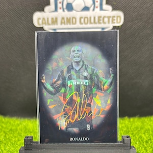 Puede incluir: Una tarjeta de intercambio holográfica negra y verde con un jugador de fútbol con una camiseta negra y verde con la palabra "IRELLI" en la parte delantera. El jugador tiene los brazos en alto. La tarjeta tiene un fondo holográfico con una firma que dice "Ronaldo" y el número "9". La tarjeta está en una caja de plástico transparente y está en un soporte negro.