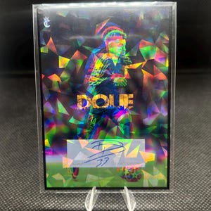 Pode incluir: Carta colecionável de futebol com o nome "DOUE" em letras coloridas. A carta apresenta um design holográfico, uma assinatura e o número "33". É apresentada numa proteção transparente, com um fundo geométrico.