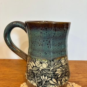 May include: A ceramic mug featuring a two-tone glaze with a dark brown rim and handle transitioning into a speckled teal upper body. The lower section displays a detailed black and white floral pattern with intricate leaves and blossoms.