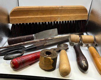 May include: A selection of antique tools, including a wooden brush, scissors, a knife, and rollers. The tools feature wooden handles and metal blades, displaying signs of use. The items are arranged on a white surface.